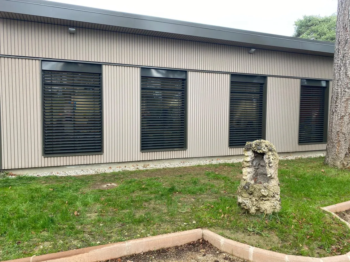 Quatre brise-soleil orientables anthracite, lames à demi ouvertes, sur les baies d’un bâtiment à bardage clair.