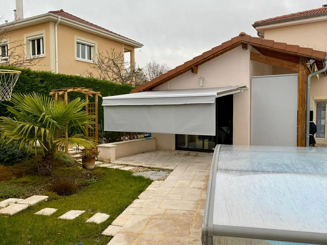 Store banne gris clair déployé au-dessus d’une terrasse dallée, son lambrequin descendu pour couper le soleil rasant.