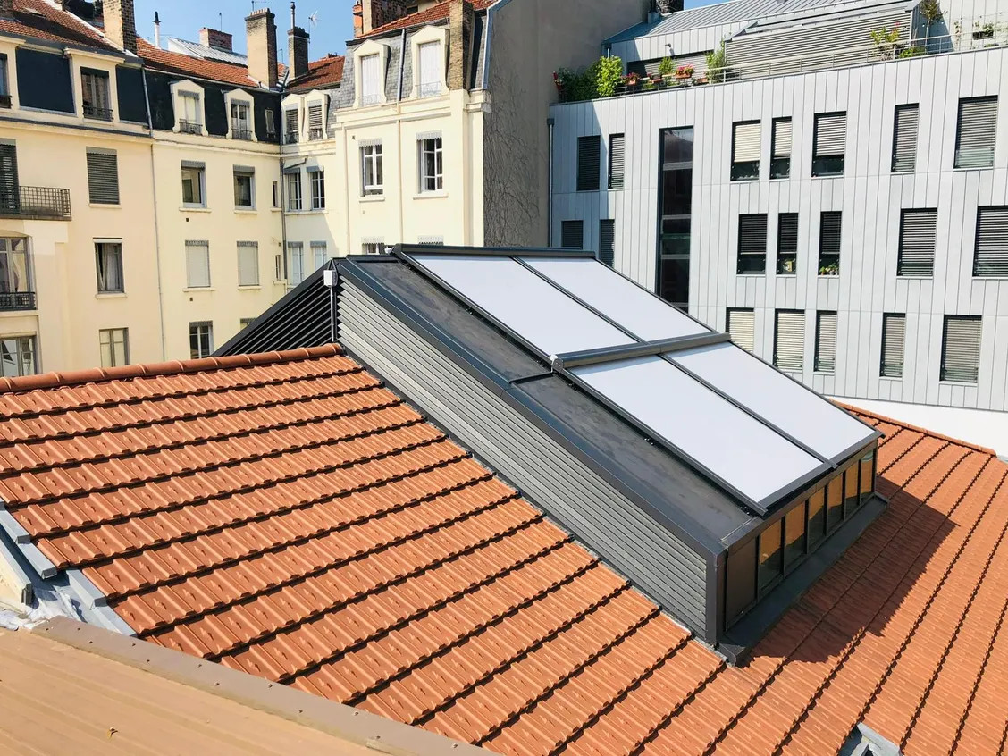 Stores de toiture à toile claire tendus sur une verrière en pente, entre un toit de tuiles et des façades d’immeubles.